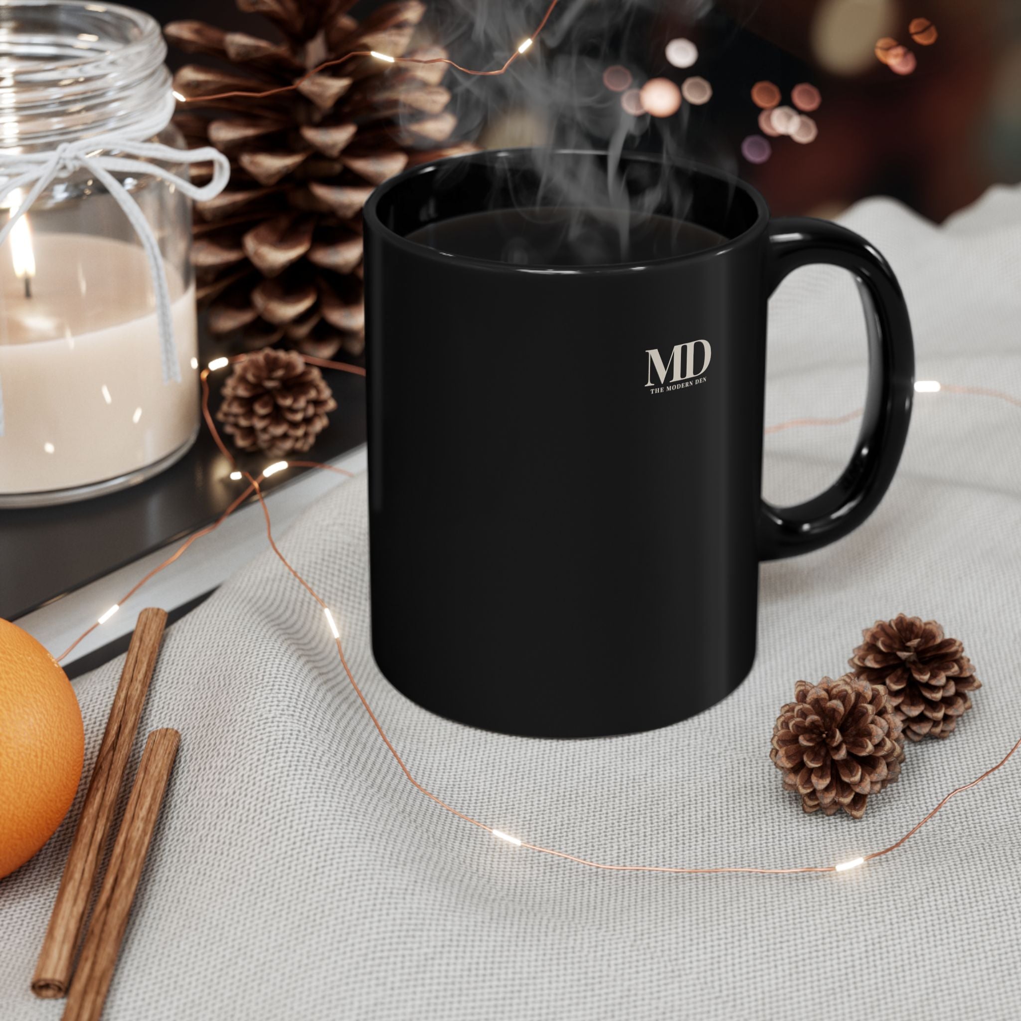 Black mug with steam, surrounded by decorative items like a candle, oranges, and pine cones on a light surface.